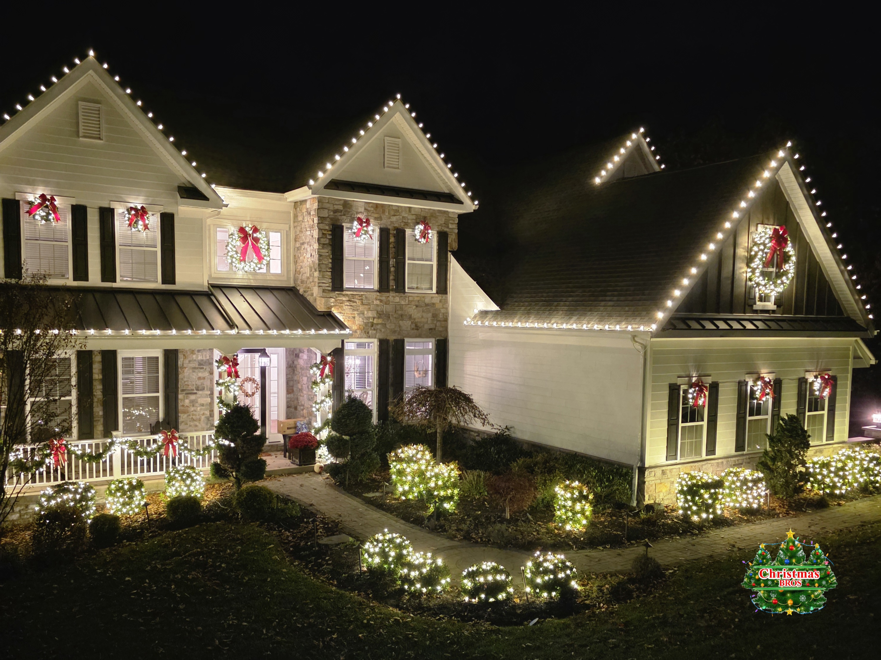 Wreaths and bows in the upper and lower windows, garland along the porch rail, a big lit wreath on the end gable, and lit bushes the whole way across the front.
