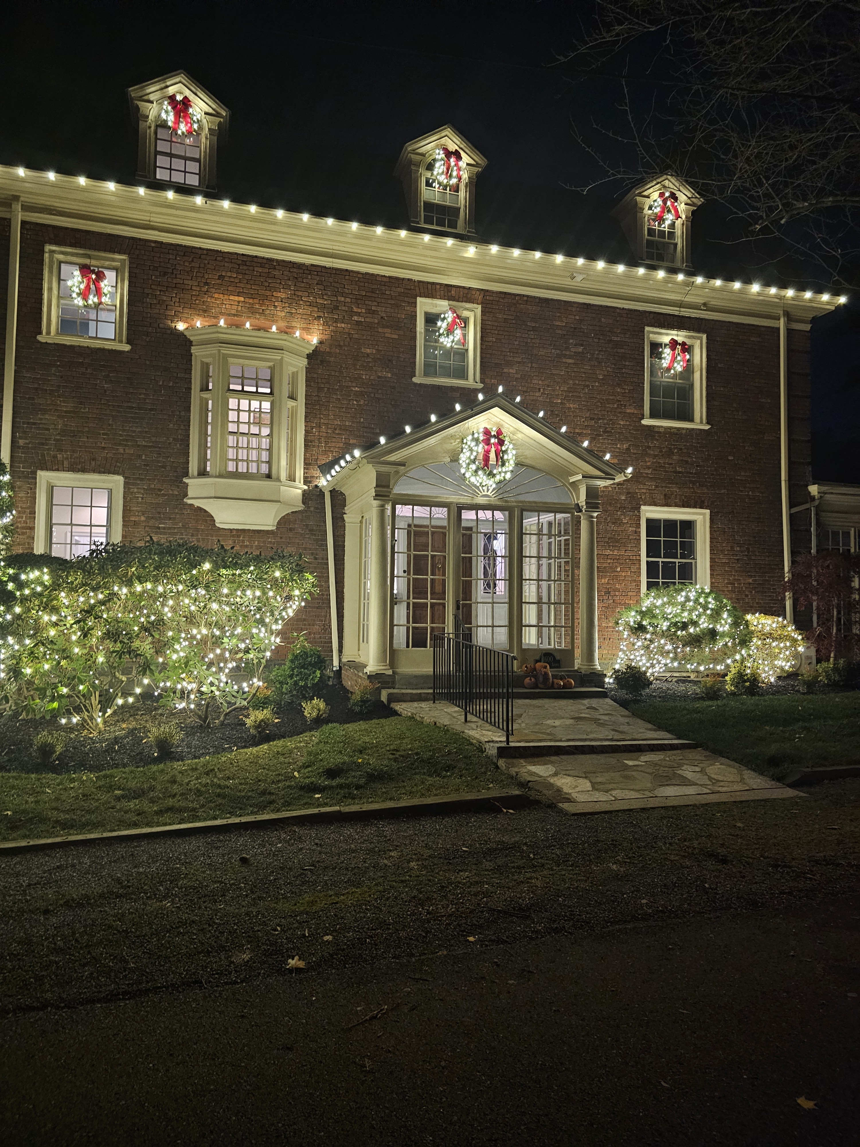Brick colonial with a wreath and bow in all three dormers, more in the windows, and a big one hung over the entry. Hedges lit right up to the walk.